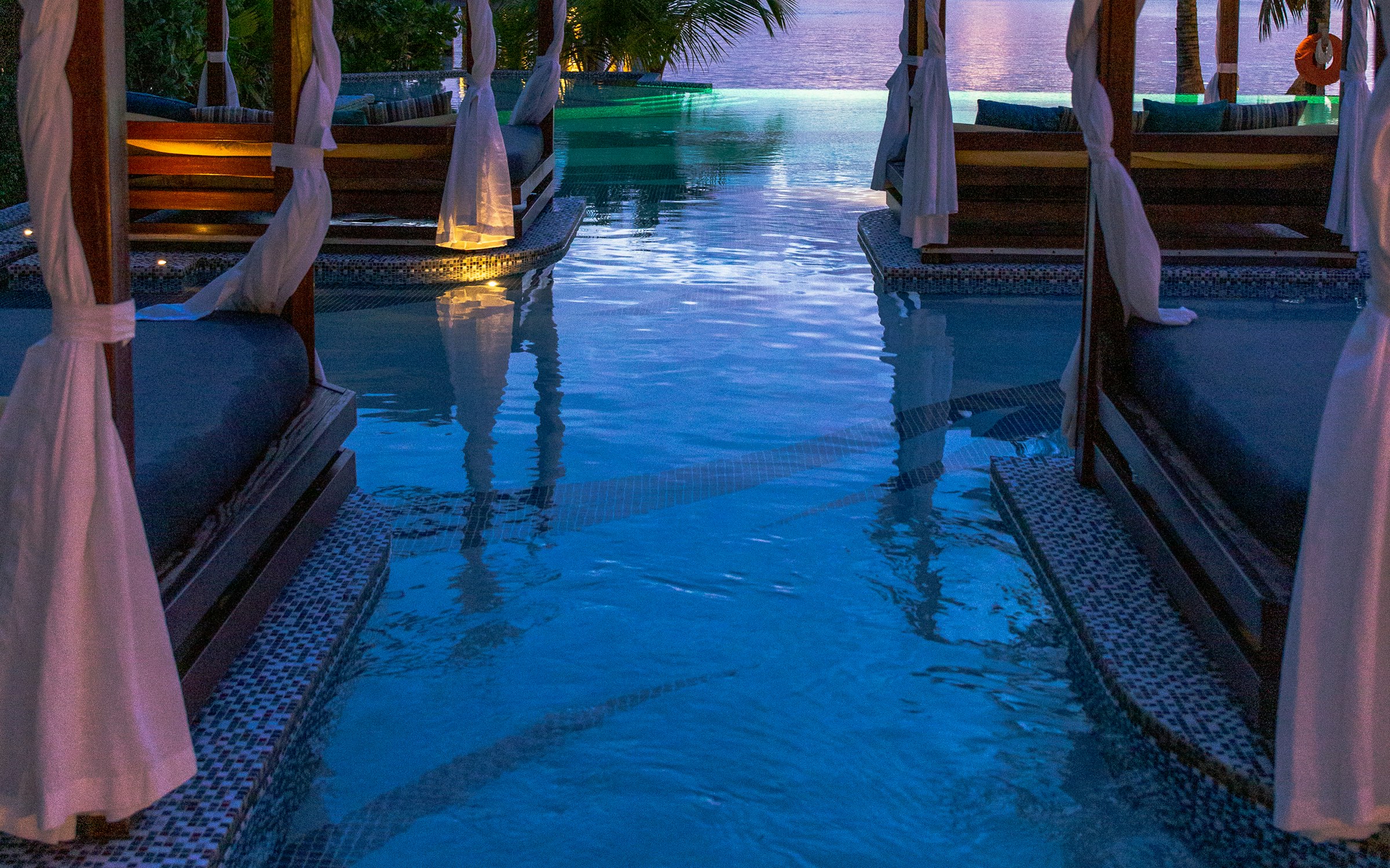 Infinity pool with shaded daybeds overlooking the ocean at dusk.